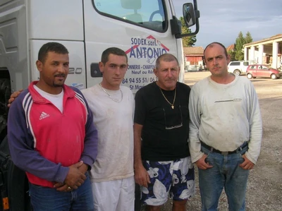 Équipe de trois ouvriers qualifiés de maçonnerie sur un chantier de rénovation de façade traditionnelle, en discussion avec le dirigeant lors d'une pause, gilets de chantier, outils de maçonnerie visibles en arrière-plan, ambiance de travail d'équipe fidèle et expérimentée.